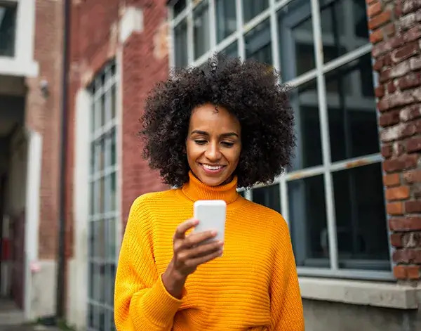 A woman with curly hair smiles brightly as she engages with her phone, radiating joy and positivity.